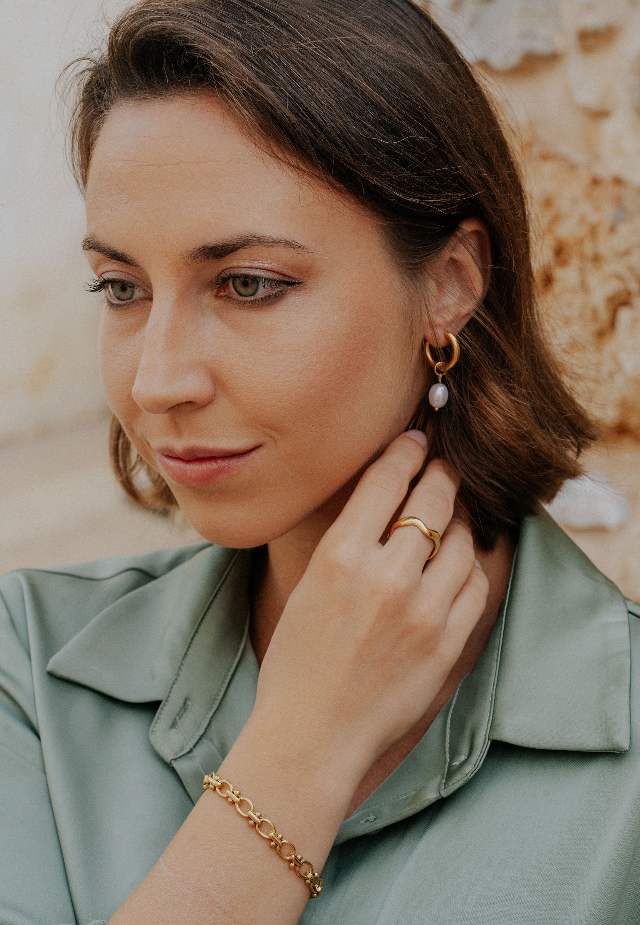 Woman adjusting an earring with a gold bracelet, wearing a light green top.