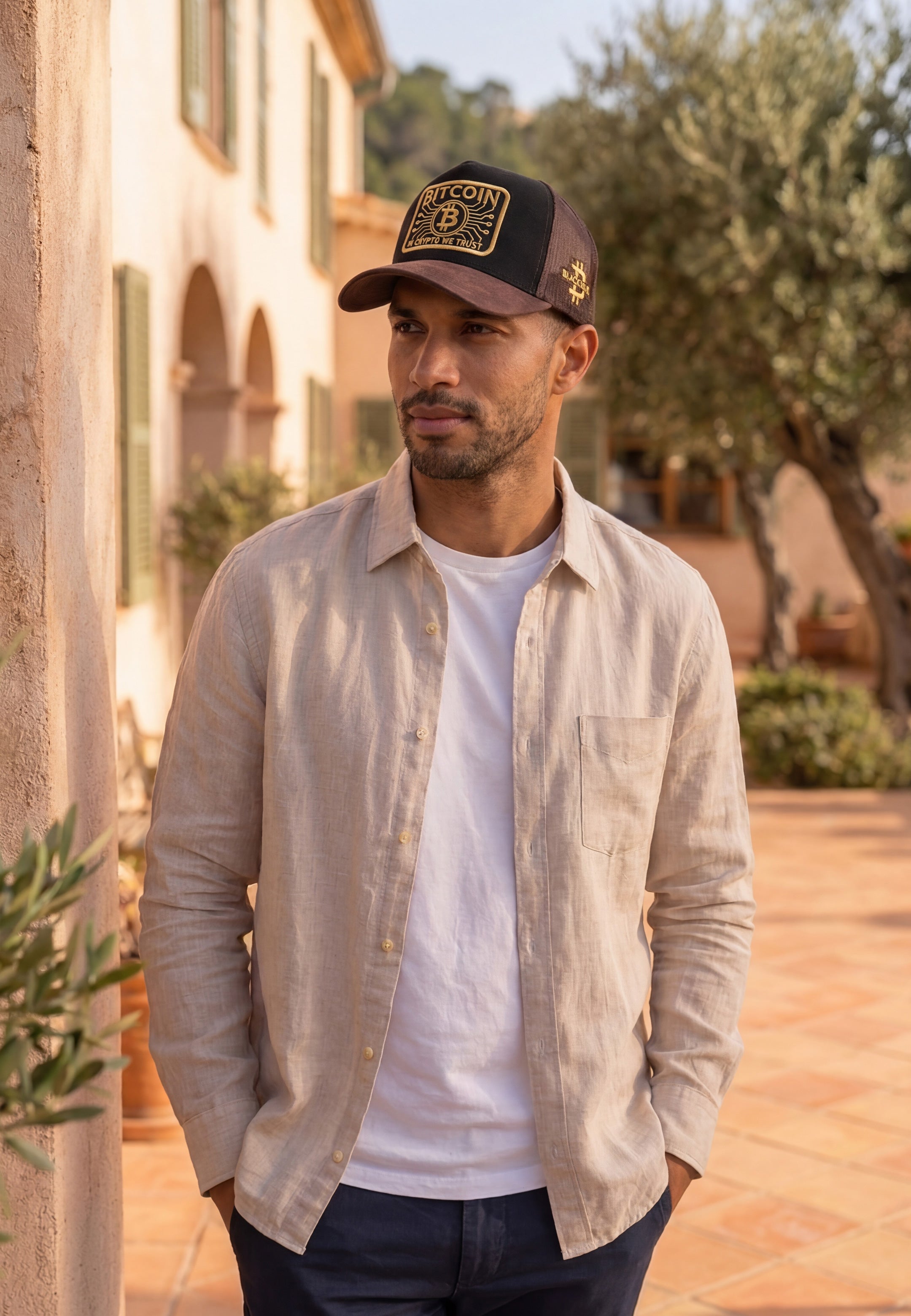 Man wearing a bitcoin cap with a logo, beige shirt, and white t-shirt outdoors.