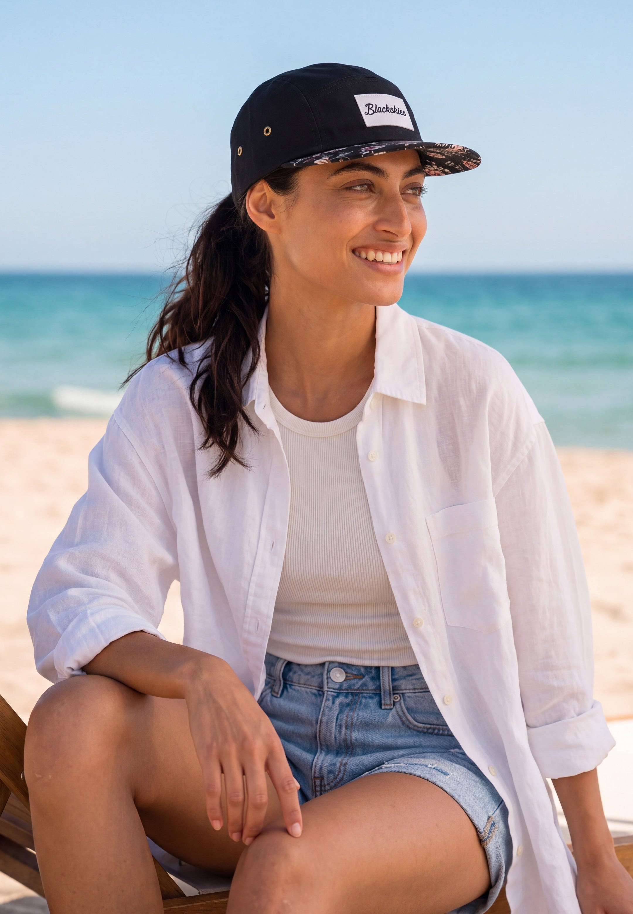 Woman sitting on a beach wearing a white shirt, denim shorts, and a black cap with text.