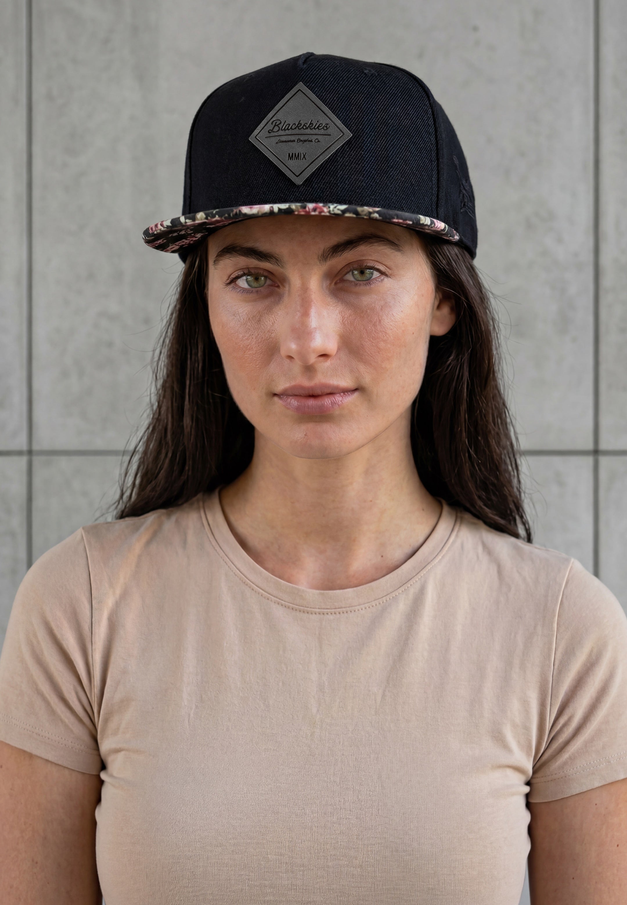 Woman wearing a black cap with a floral pattern on the brim against a gray tiled wall.