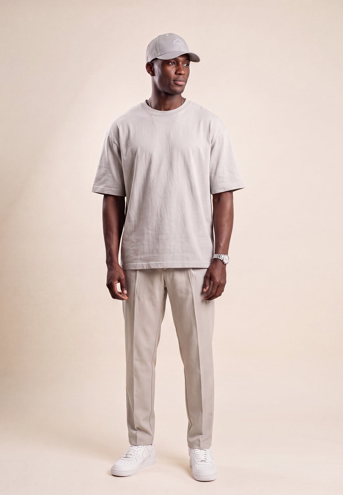 A man modeling a light gray crewneck t-shirt, beige trousers, white sneakers, and a gray baseball cap against a solid beige studio background.