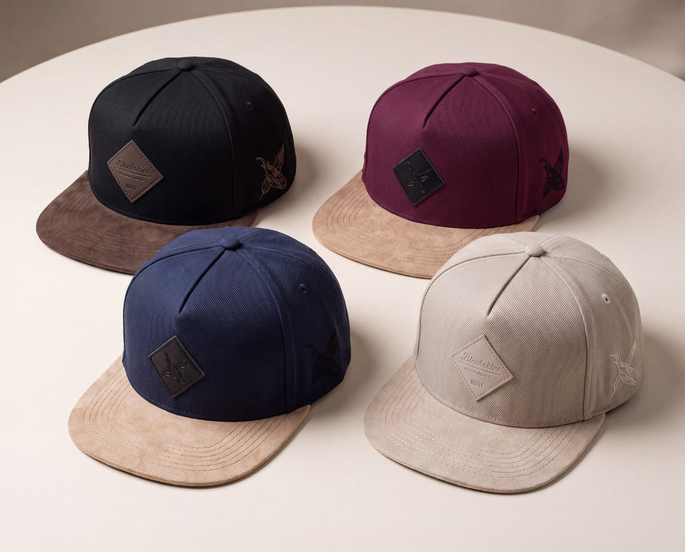 Four snapback cap hats arranged on a round table, each with a structured fabric crown, a flat brim, and a small diamond-shaped patch on the front. The colors shown are black with brown, burgundy with tan, navy with tan, and light gray with tan.