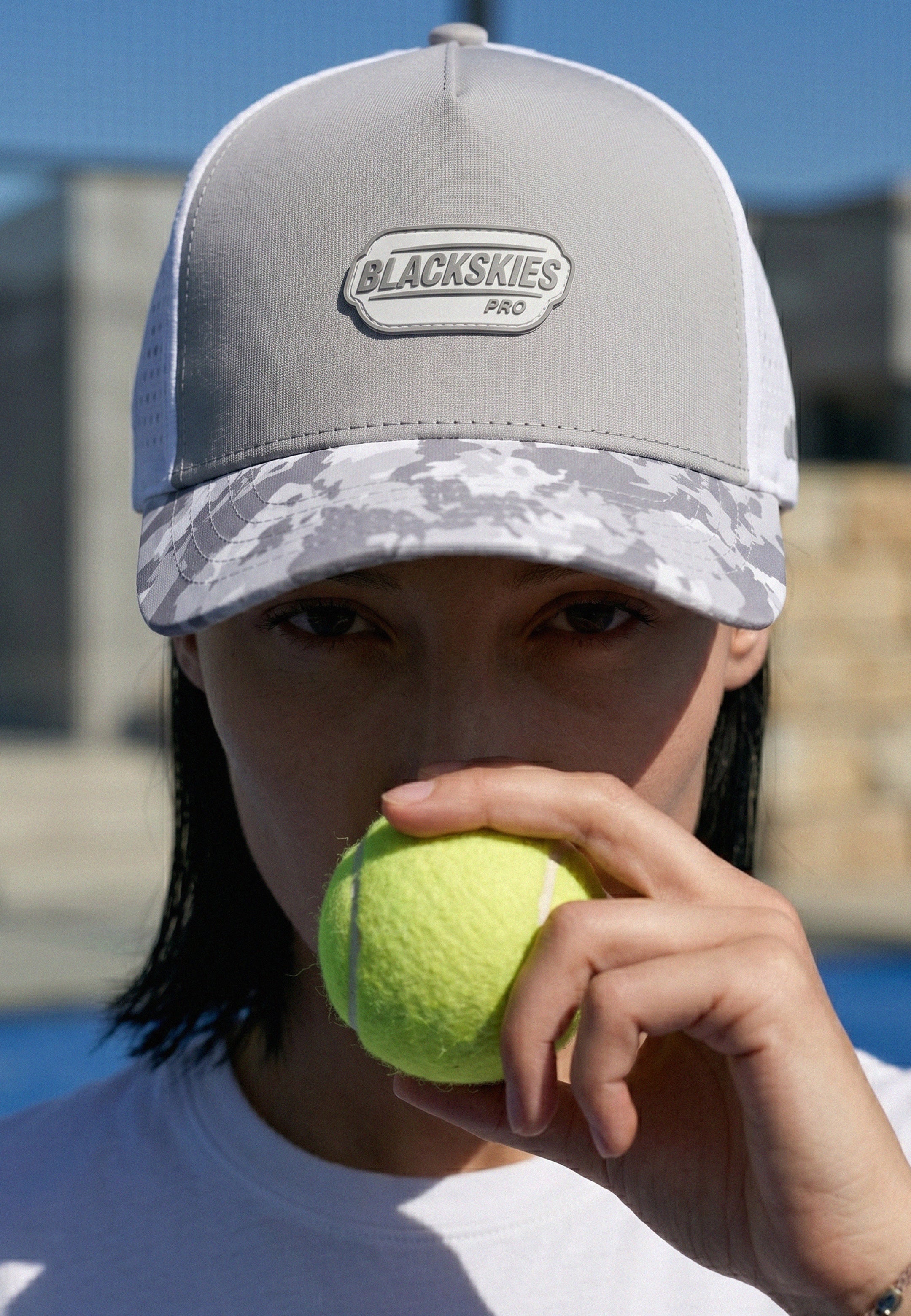 Person holding a tennis ball with a 'Blackskies' cap on a sports court