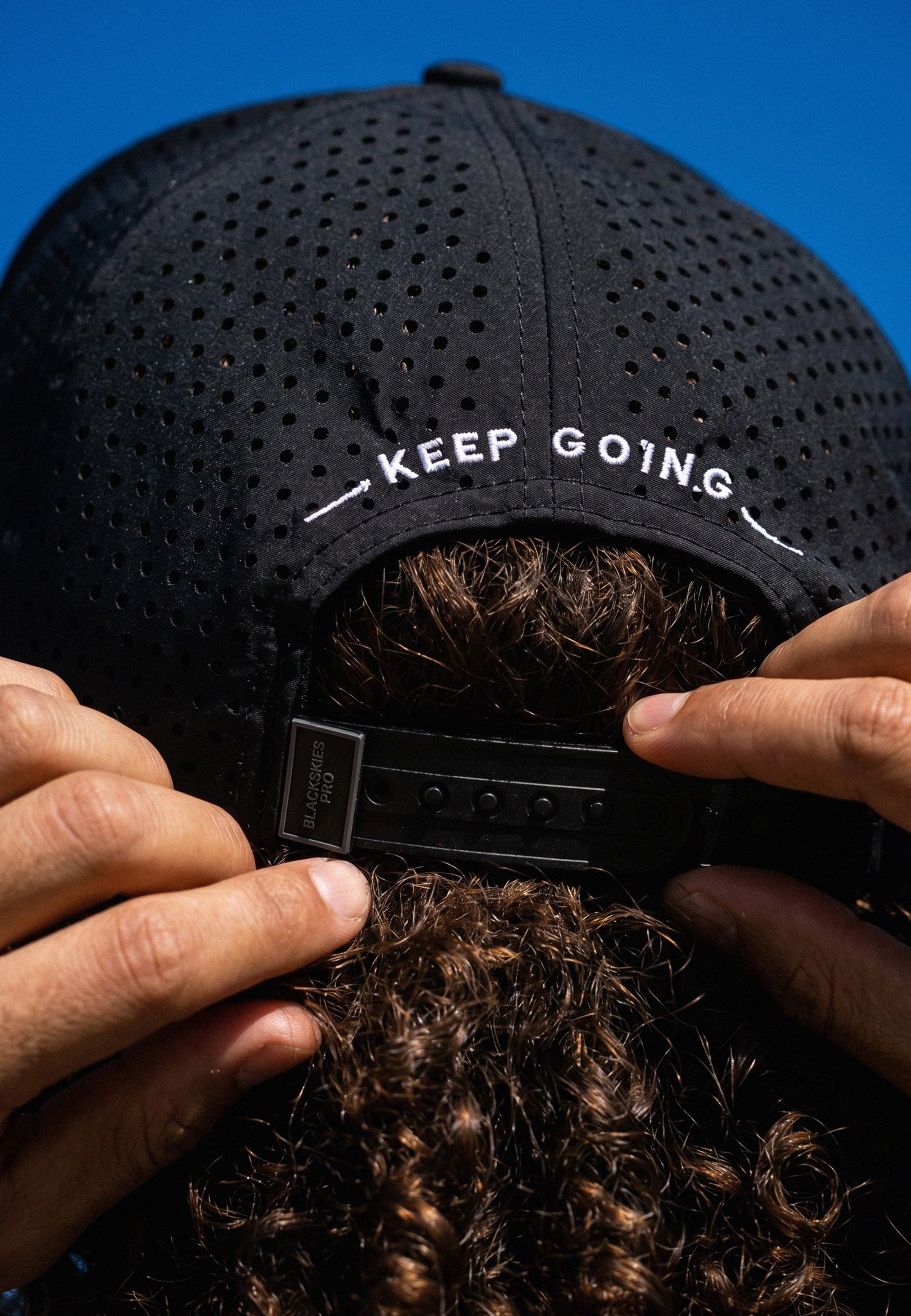 Black cap with 'KEEP GOING' text held by a person against a blue background