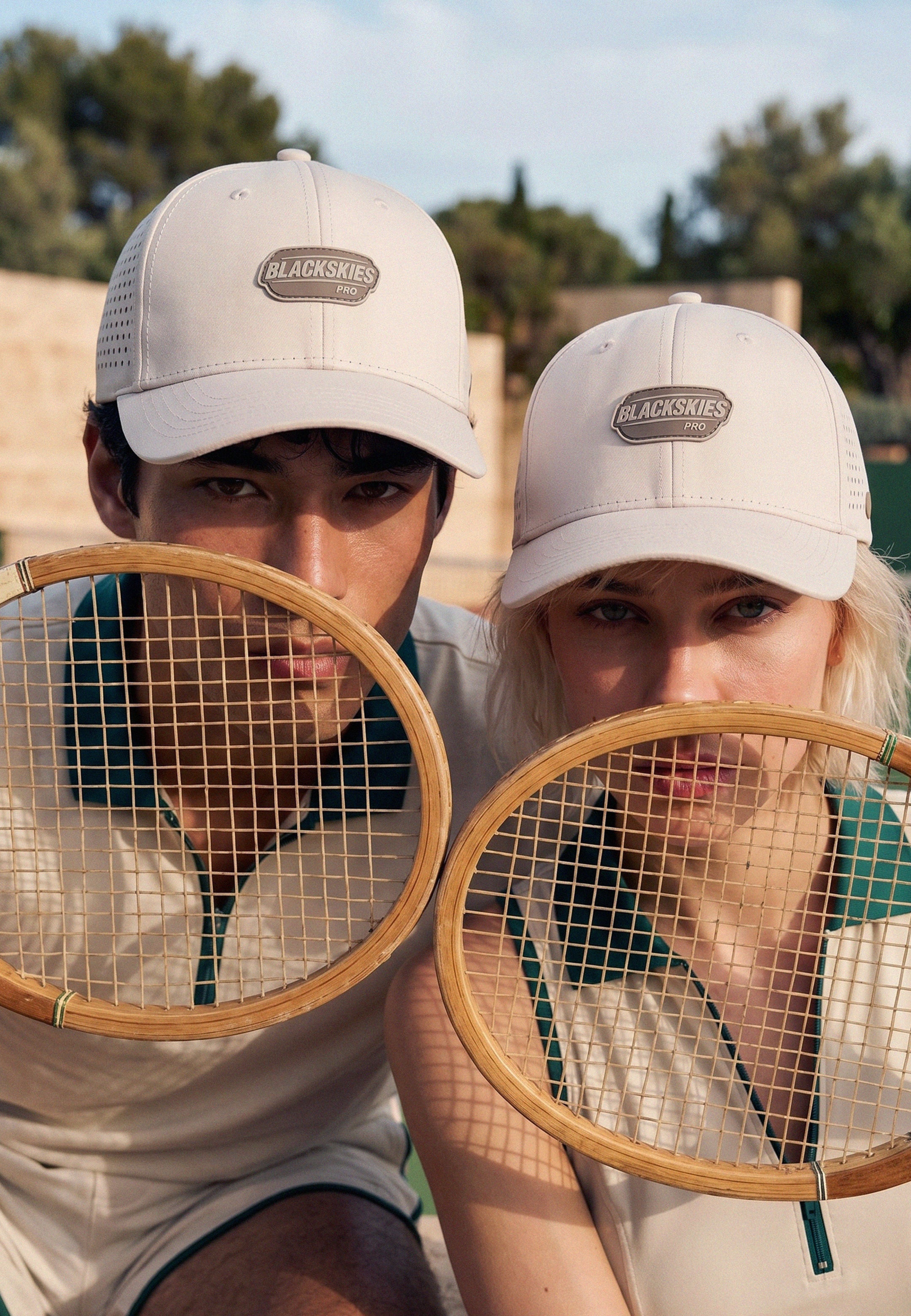 Two people on a tennis court wearing vintage-style clothing and holding wooden tennis rackets.