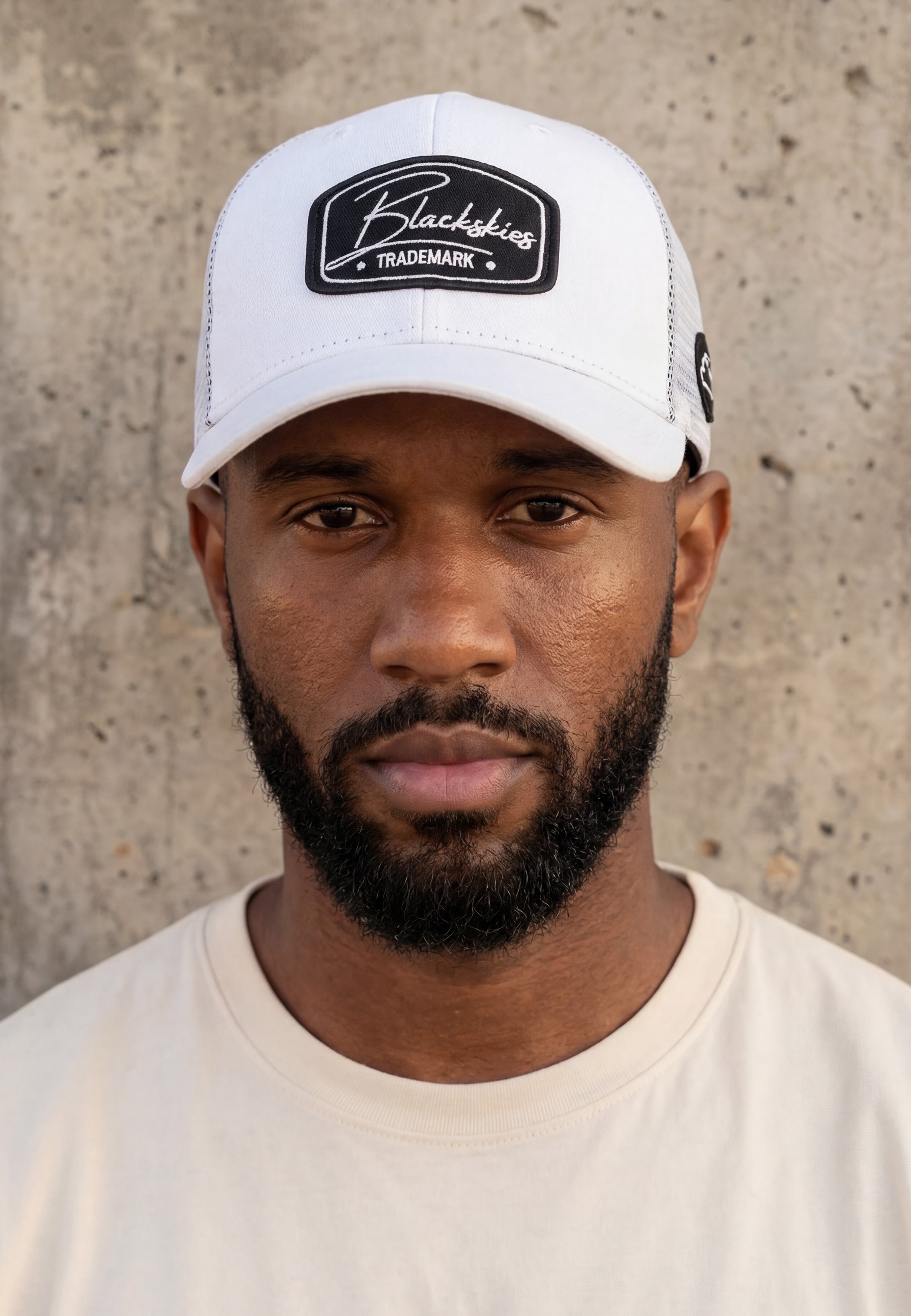 Man wearing a white cap with a blackskies brand logo against a concrete wall.
