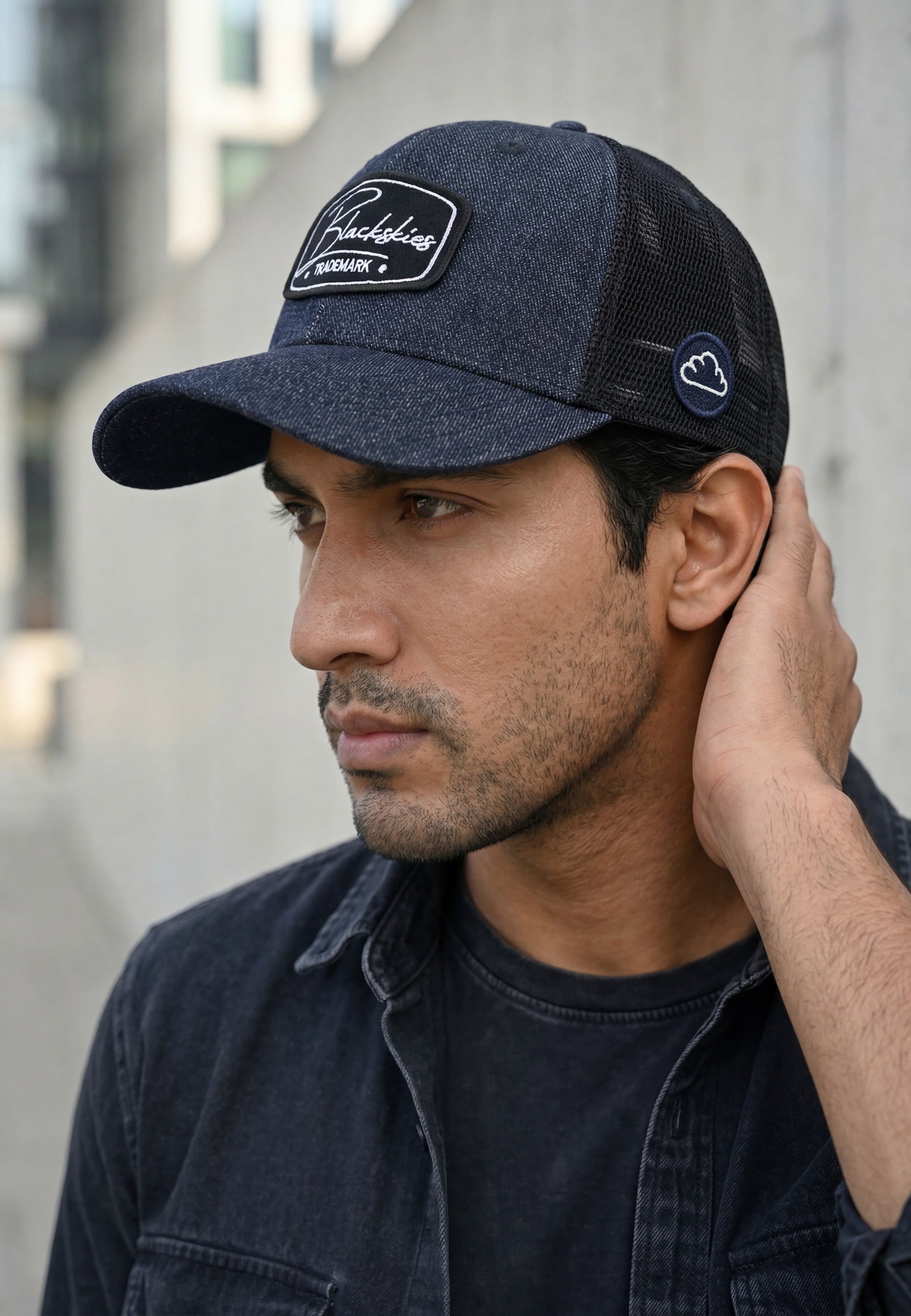 Man wearing a navy blue denim trucker cap with a blackskies  logo, adjusting it with his hand.