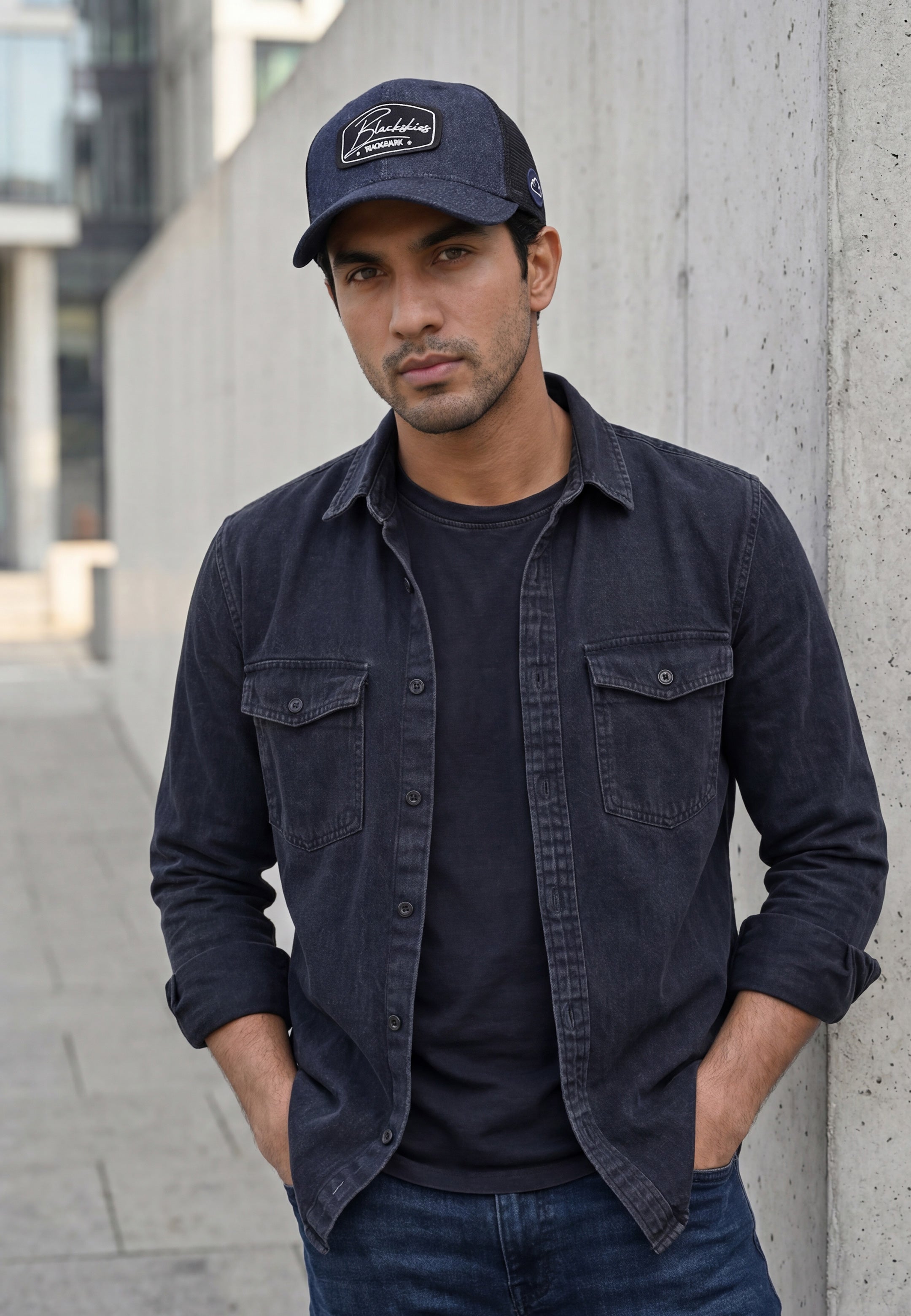 Man wearing a dark denim jacket, blue denim cap, and jeans standing against a concrete wall.