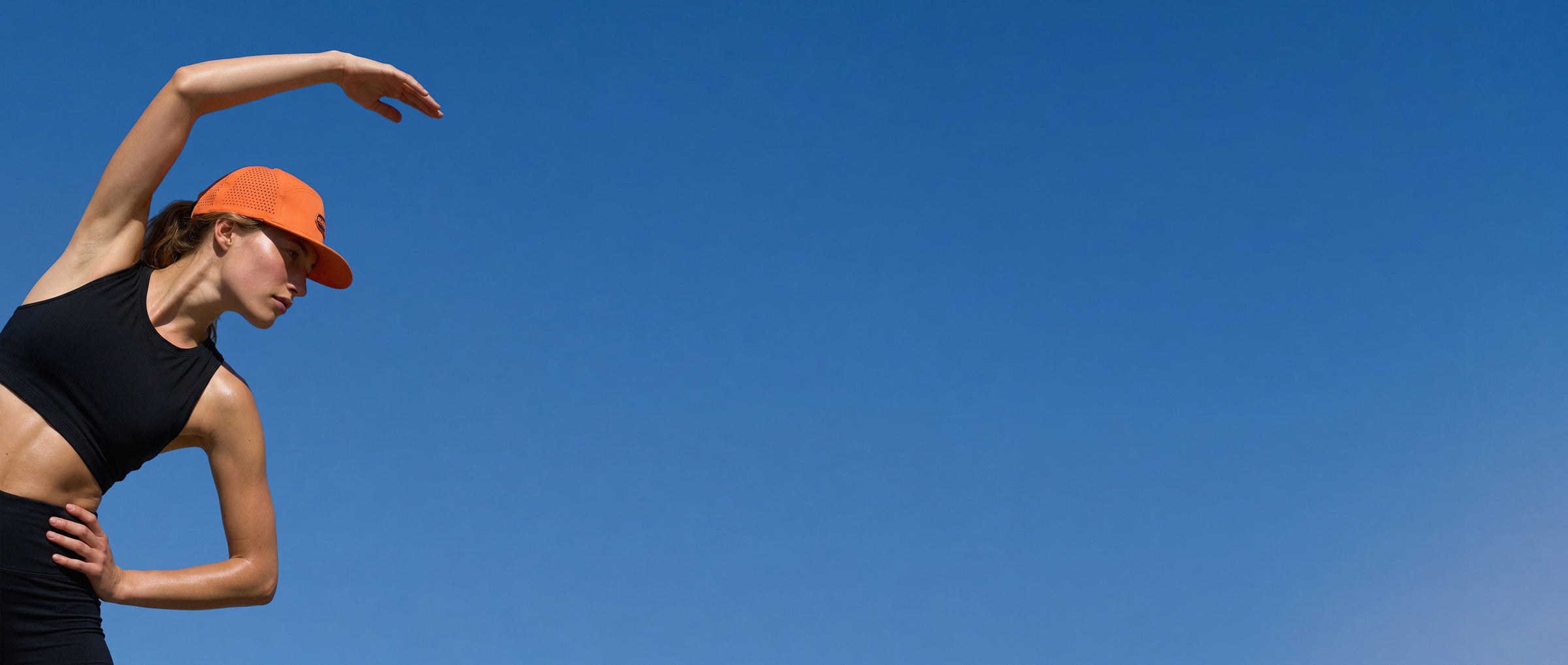 Woman in black sports bra and orange cap stretching against a clear blue sky